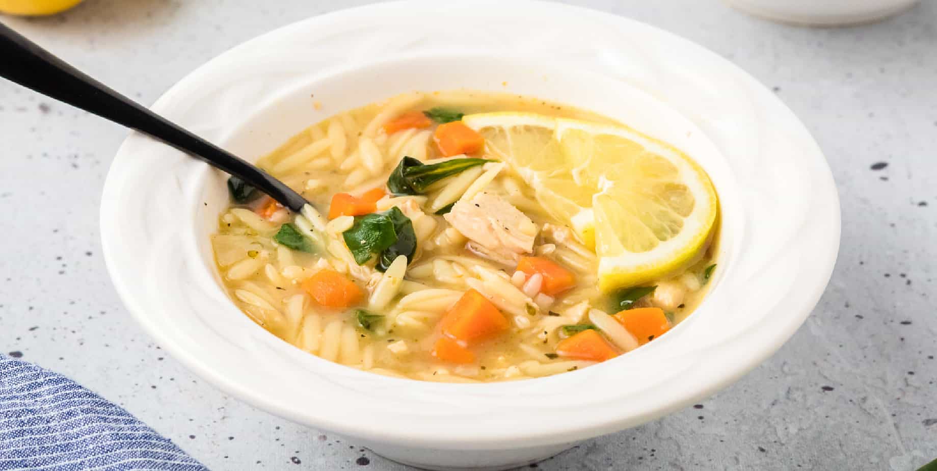 Wide view of a bowl of soup with chicken, carrots, spinach, orzo pasta and lemon in the bowl.