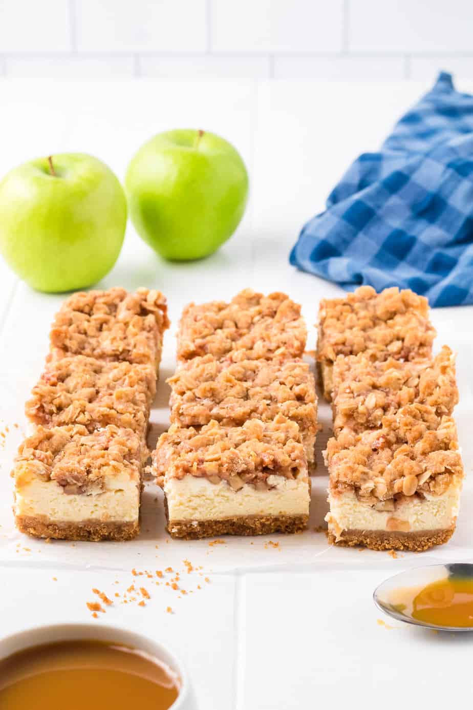 Sliced apple cheesecake bars on a counter with apples and caramel nearby on the counter.