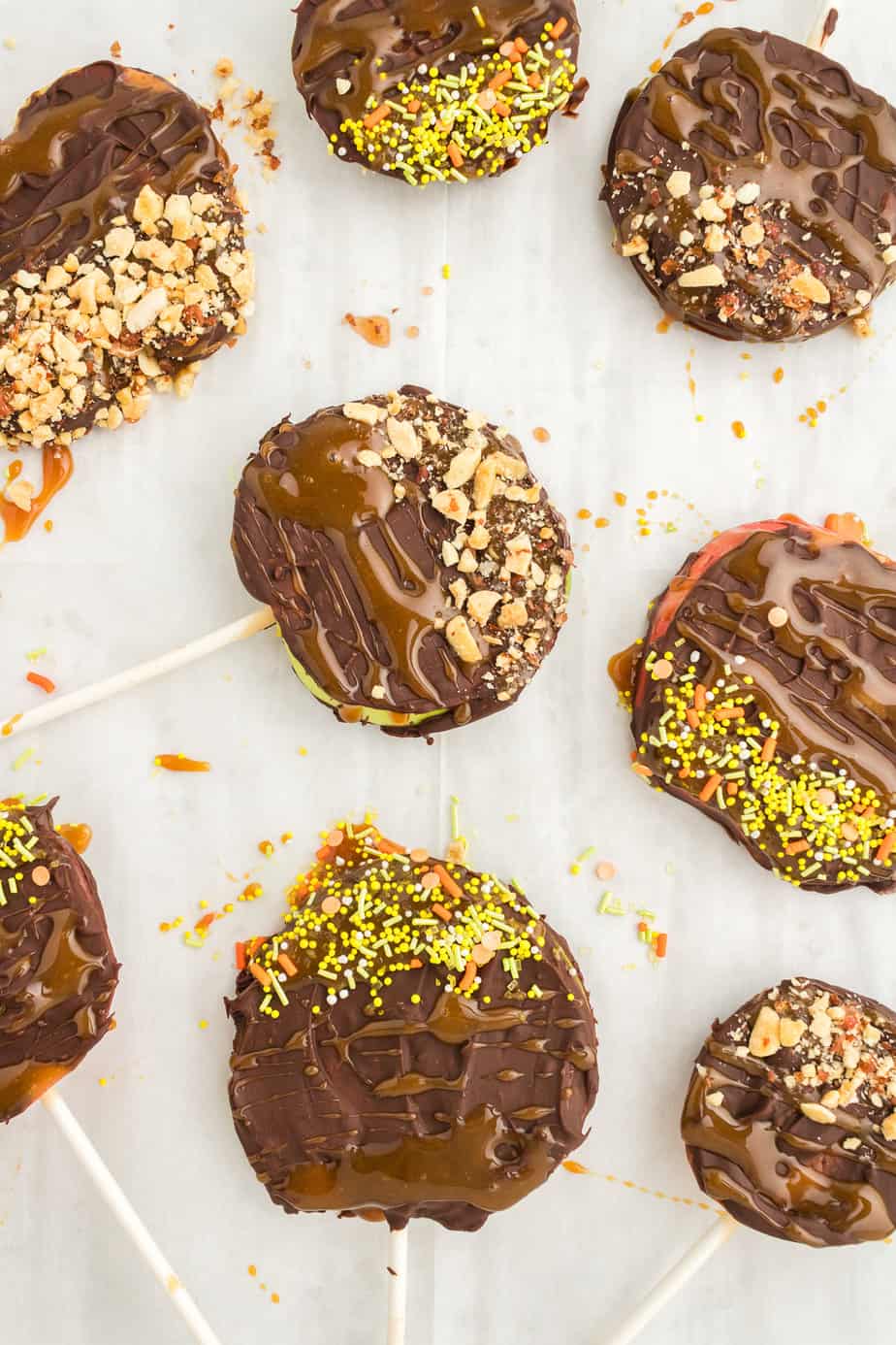 Apple slices with sticks coated in chocolate, caramel nuts and sprinkles from overhead close up on parchment paper.