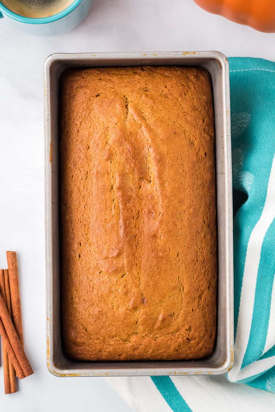 Baked pumpkin bread in a loaf pan from overhead on a counter with a linen and cinnamon next to the pan along the edges.