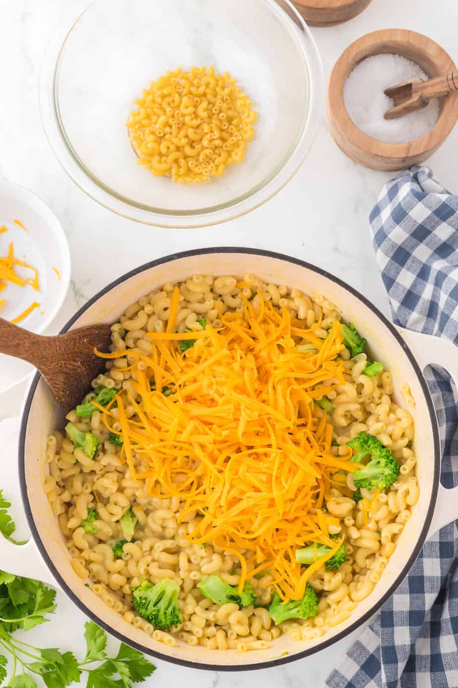 Shredded cheddar cheese being stirred into a pot of cooked macaroni and broccoli in a pot from overhead with more ingredients in bowls on the counter nearby.