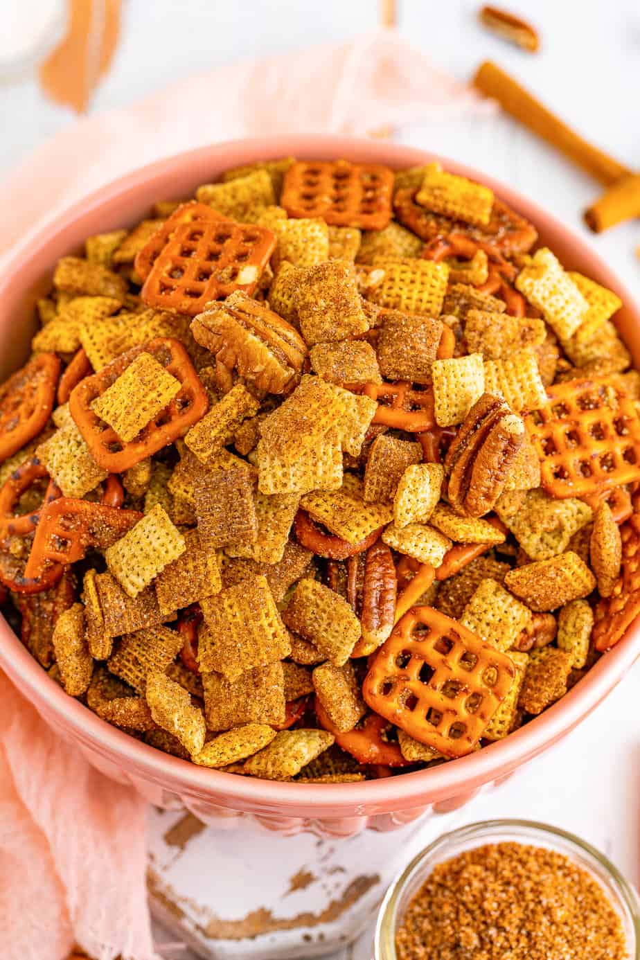 Close up of cinnamon sugar chex mix in a large serving bowl from overhead on a counter with some of the crunch topping, cinnamon sticks and a linen nearby on the counter.