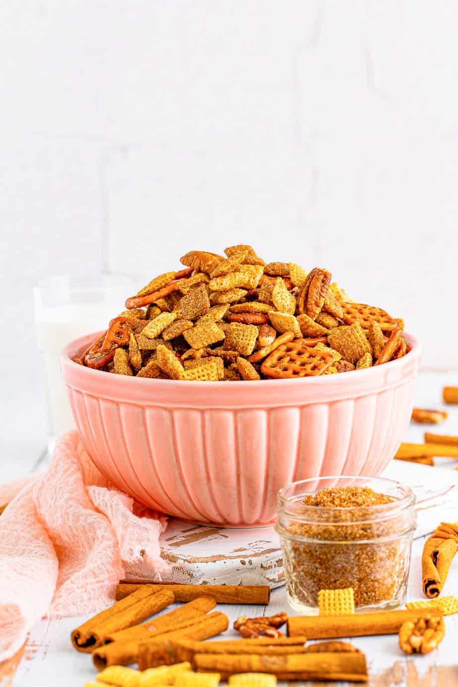 Finished cinnamon sugar chex mix in a large pink bowl piled high from the side with more cinnamon and more of the crunch topping in a container nearby.