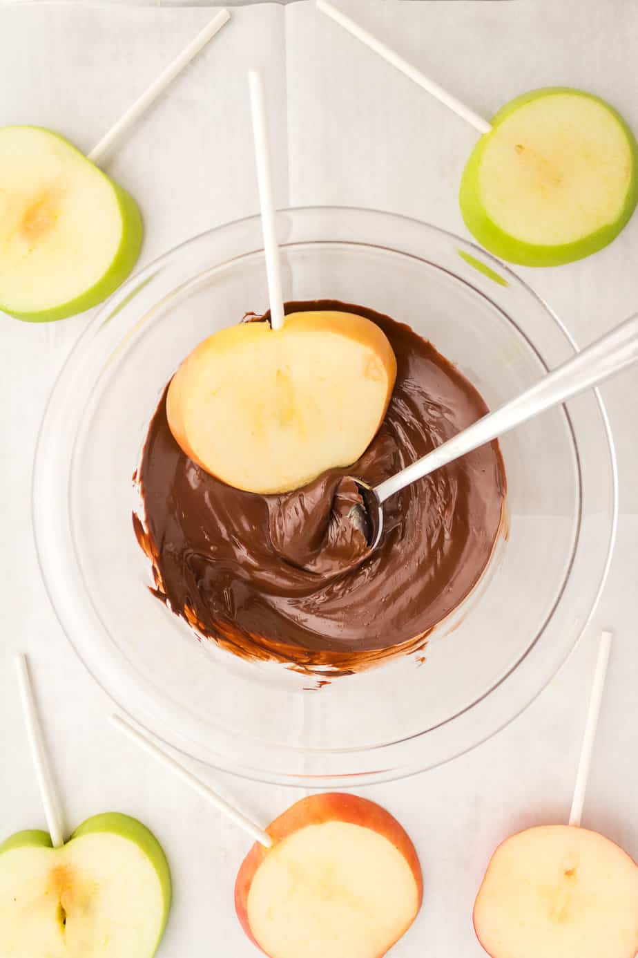 Dipping an apple slice with a stick in a bowl of chocolate with a spoon from overhead with more apple slices on the counter nearby.