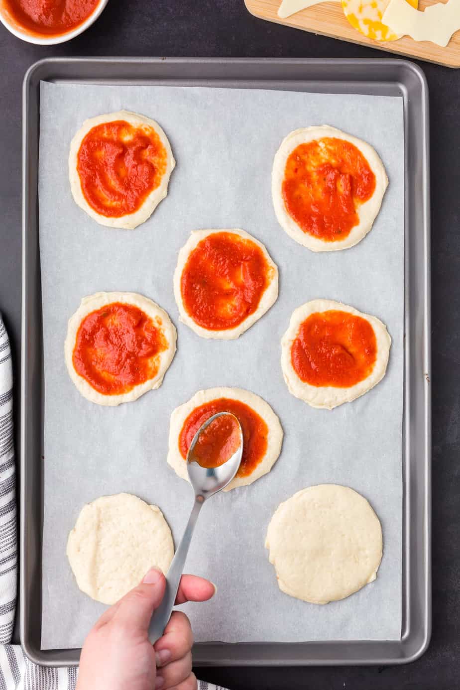 Adding tomato sauce with a spoon to each mini pizza crust spaced out on a baking sheet.