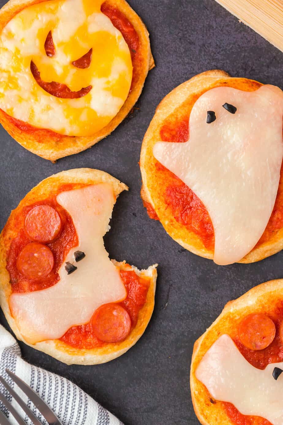 Halloween mini pizzas decorated with cheese shaped like a bat, ghost and jack-o-lantern up close with the bat missing a bite.