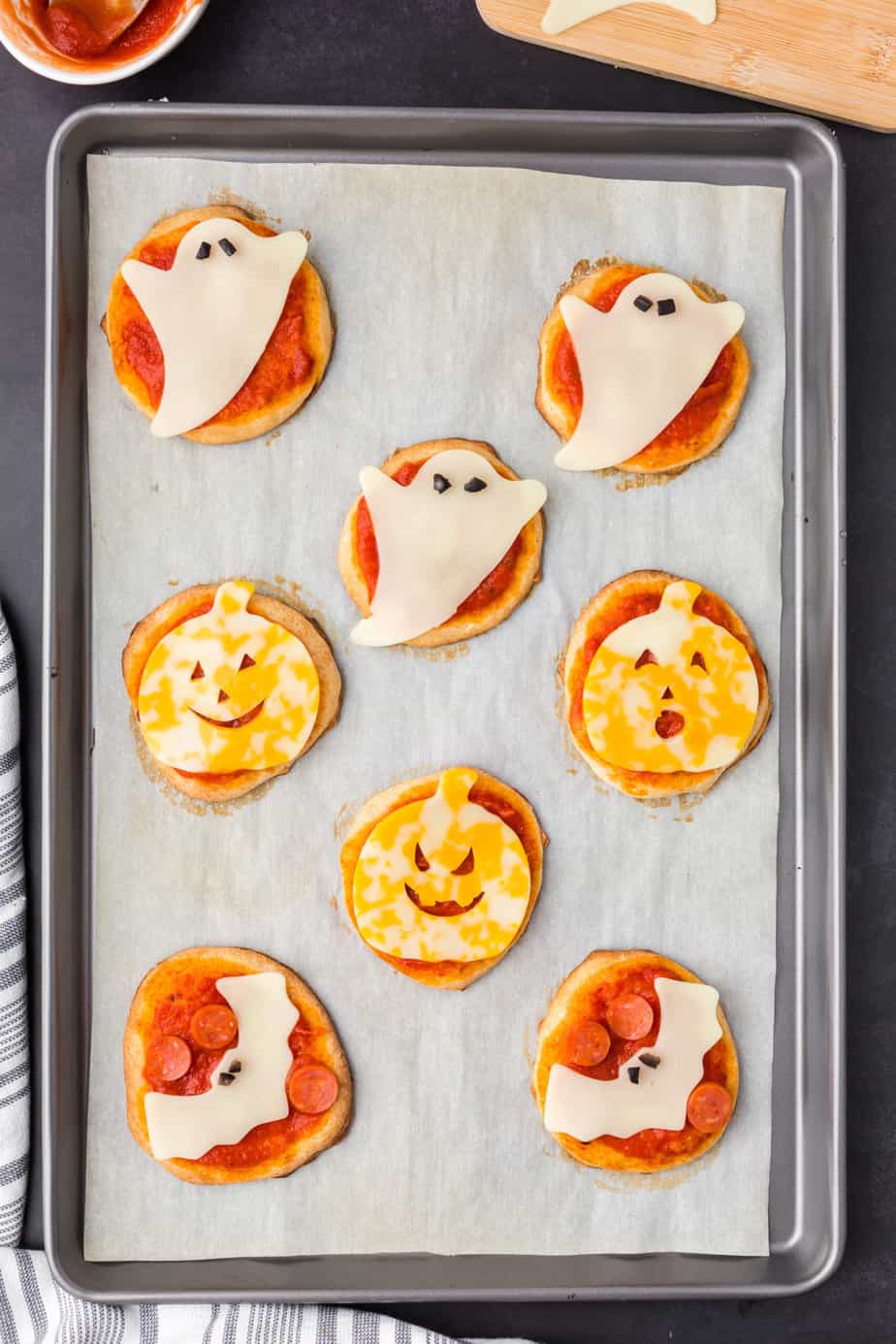 Adding pumpkin, ghost and bat shaped cheese with olive eyes to mini pizzas on a baking sheet.