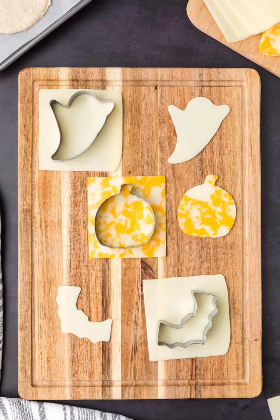 Pumpkin, ghost and bat cookie cutters cutting cheese slices on a cutting board.