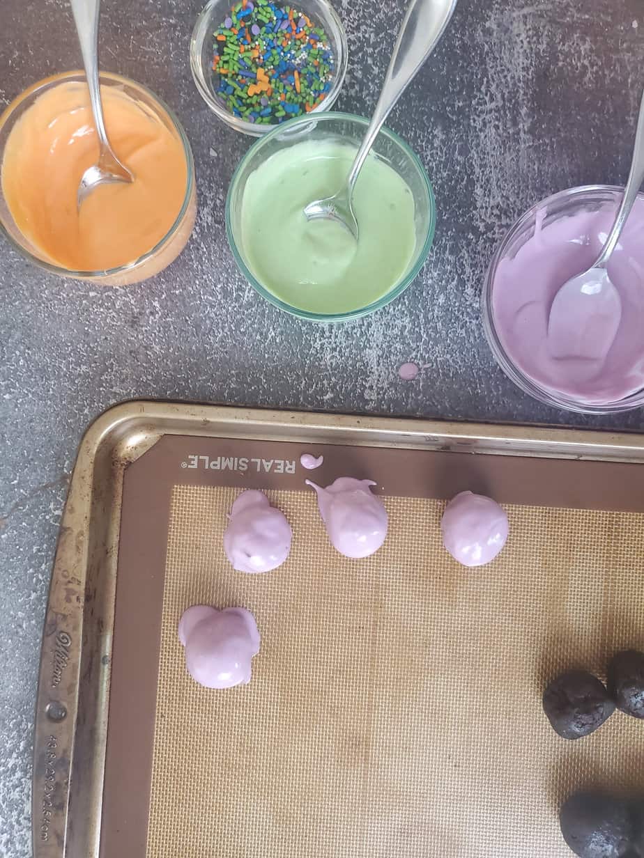 A bowl of melted orange, green and purple chocolate wiith spoons on a counter next to a pan holding oreo truffles that have been dipped in purple chocolate from overhead.