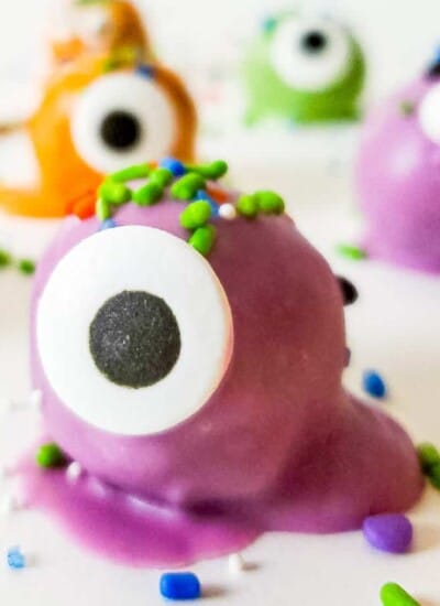 Close up side view of a purple coated truffle decorated with sprinkles and a candy eye with more monster truffles dipped in orange, green and purple chocolate in the background.
