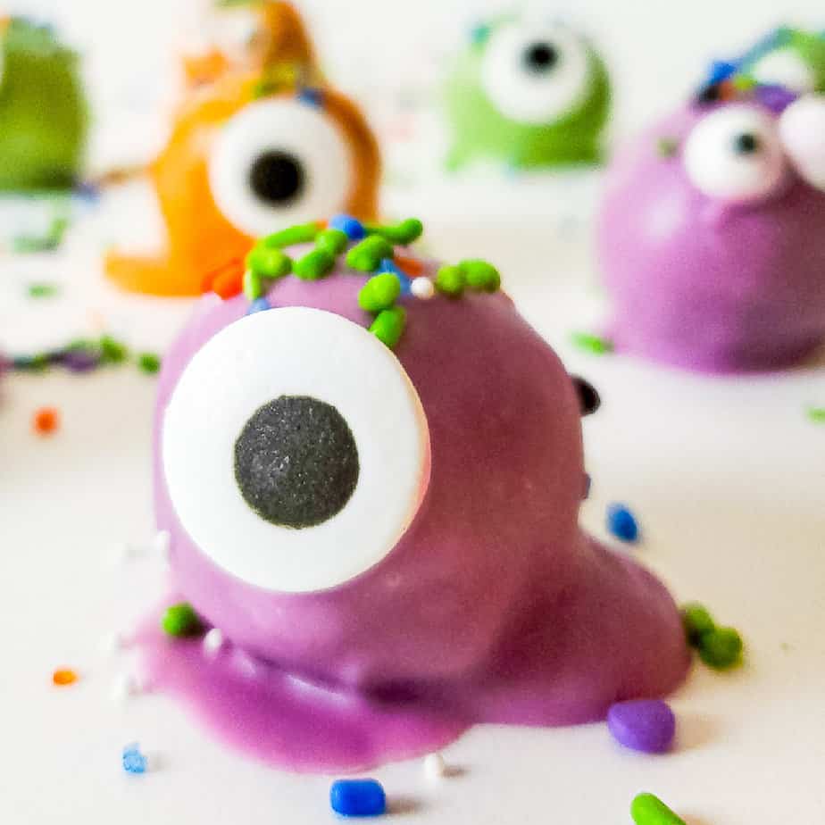 Close up side view of a purple coated truffle decorated with sprinkles and a candy eye with more monster truffles dipped in orange, green and purple chocolate in the background.