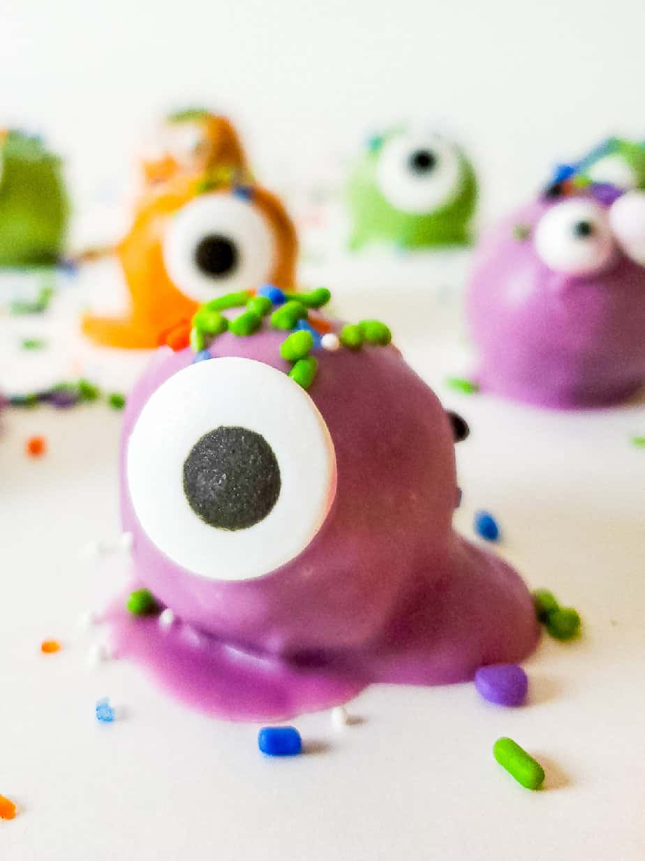 Purple oreo ball from the side decorated with a large candy eye and sprinkles to look like a monster with more orange, green and purple chocolate truffles behind decorated like monsters.