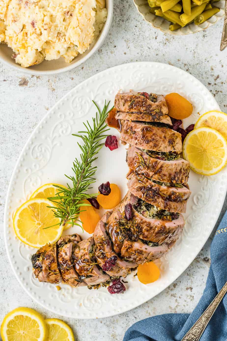 Pork tenderloin stuffed and sliced into rounds on a serving platter from overhead on a kitchen counter. Slices of lemon, rosemary and dried fruit garnish the platter and sides of green beans and mashed potatoes can be seen at the edge of the image.