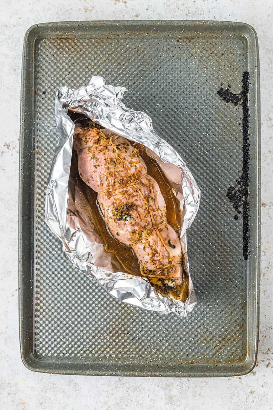 Stuffed and rolled pork tenderloin sits in a piece of foil rolled up on the sides to contain the glaze on top of a baking sheet after cooking.