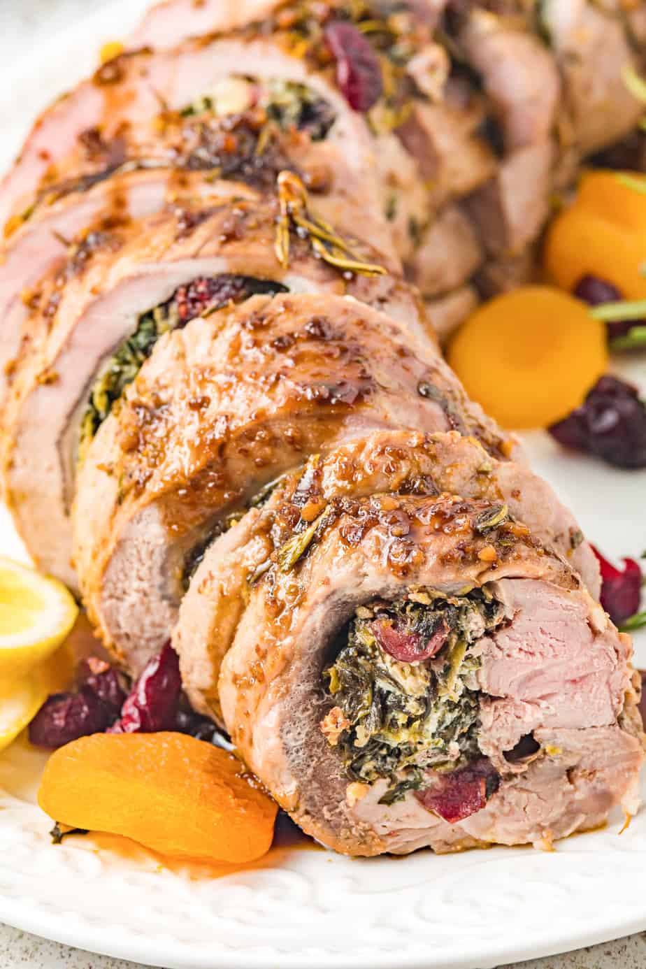 Close up stuffed pork tenderloin on a platter from the side with dried apricots and cranberries on the plate nearby and the pork sliced into portions on a serving platter.