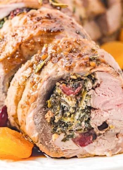 Close up of rolled pork tenderloin stuffed with spinach, cheese and dried fruit close up from the side with a few dried apricots and cranberries around the meat on the plate.