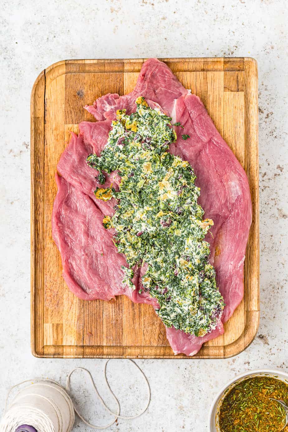 Pork tenderloin on a cutting board butterflied flat with spinach cheese filling spread over top and m ore ingredients nearby on the kitchen counter from overhead.