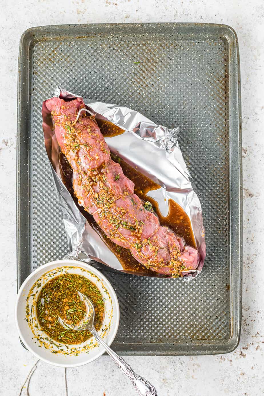 Pork tenderloin that has been rolled, stuffed and tied on top of a piece of aluminum foil with the sides pulled up on a baking pan with glaze spooned over the pork from overhead on a counter..