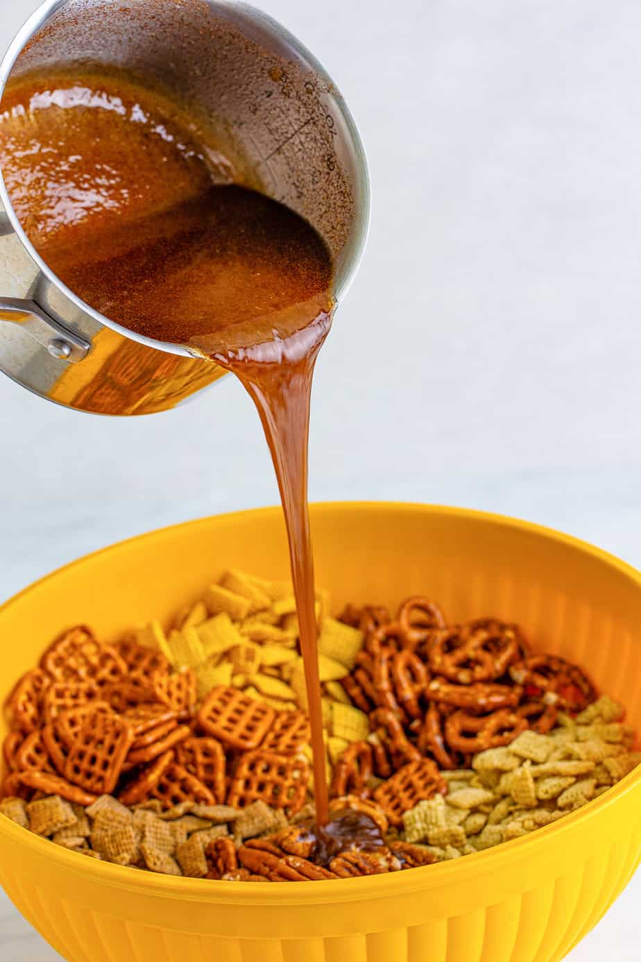 Pouring the brown sugar butter mixture from a saucepan over the chex mix in a large bowl from the side.