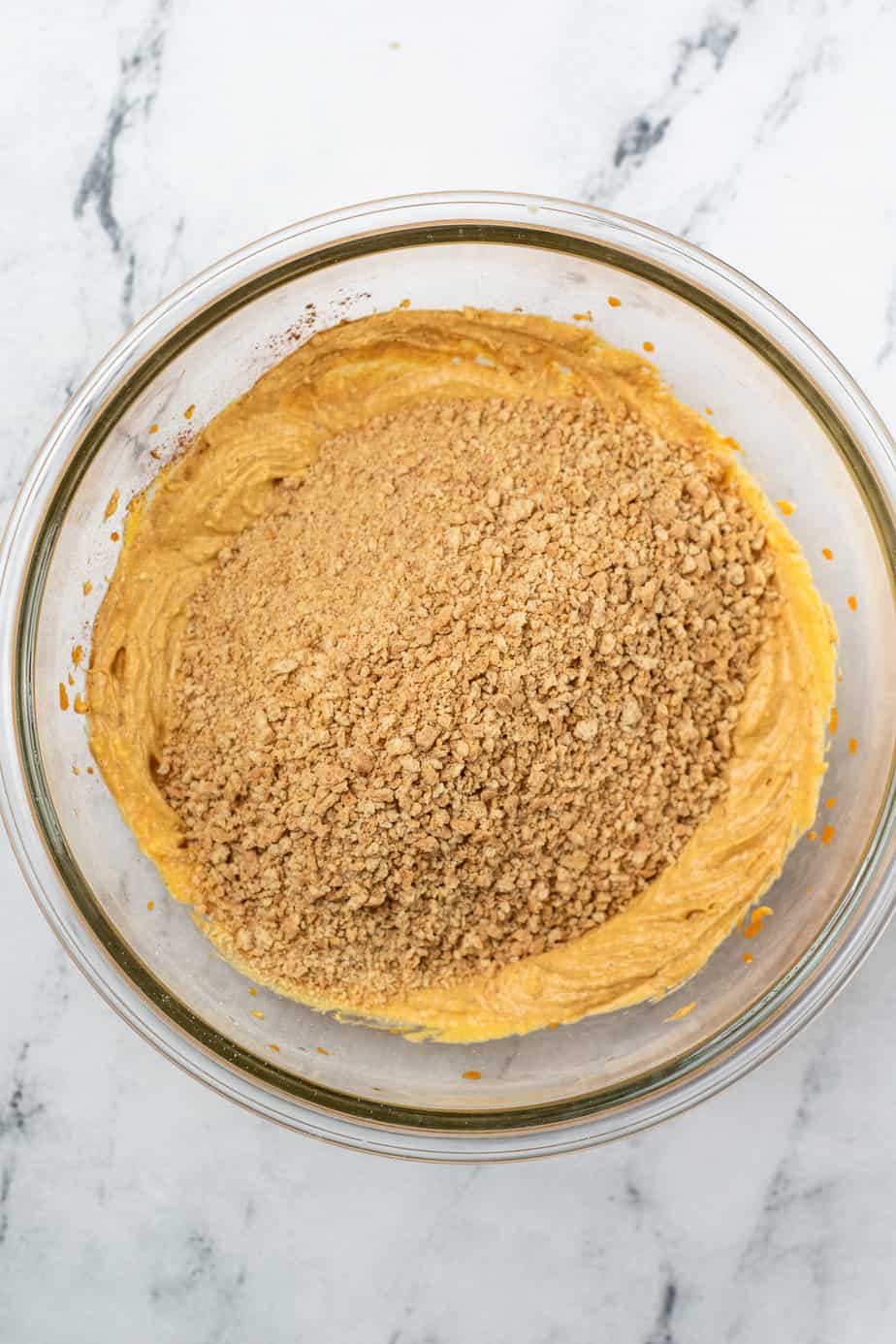 Graham cracker crumbs poured in a large mixing bowl on top of pumpkin cheesecake mixture to be blended on a counter from overhead.