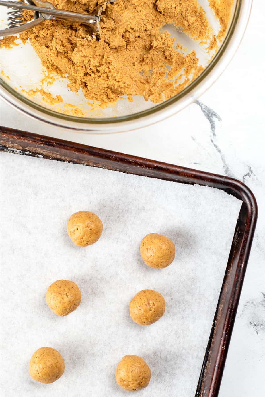Pumpkin cheesecake dough in a bowl with a cookie scoop from overhead next to a baking sheet lined with parchment paper and six pumpkin truffles placed on the pan with an inch of space between each truffle