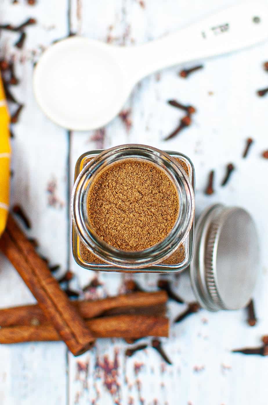 Open glass jar of pumpkin pie spice from overhead with spices, measuring spoons and sticks of cinnamon on the table nearby.