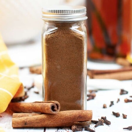 Pumpkin pie spice in a glass spice jar with a metal closed lid from the side with cinnamon and cloves on the counter in the background.