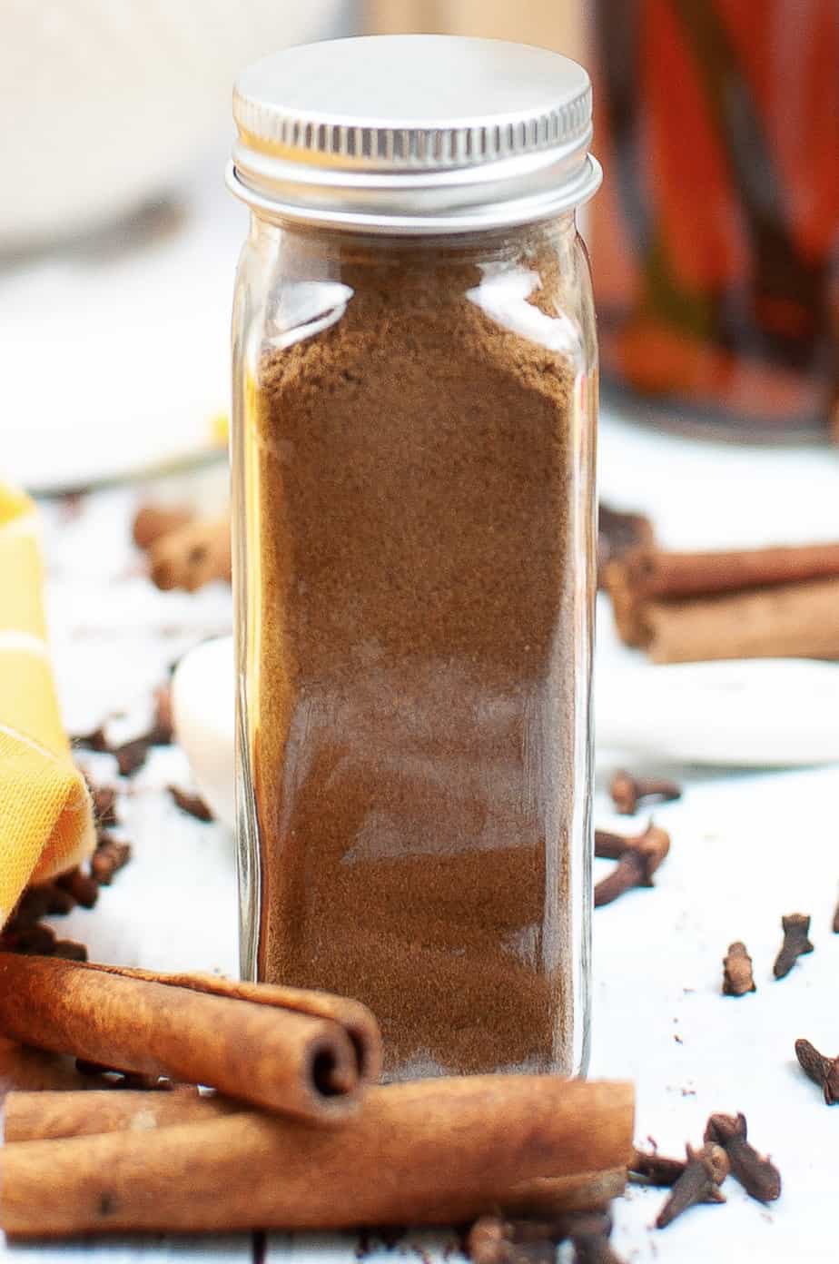 Pumpkin pie spice in a clear jar with a metal lid from the side with cinnamon next to it on the table.
