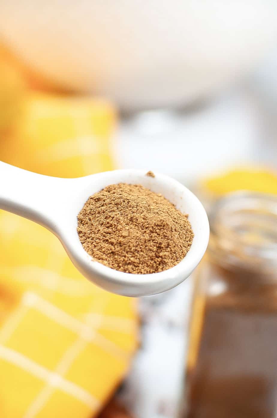 Pumpkin pie spice mix in a measuring spoon up close with a jar of spice mix and a yellow linen in the background on the table.