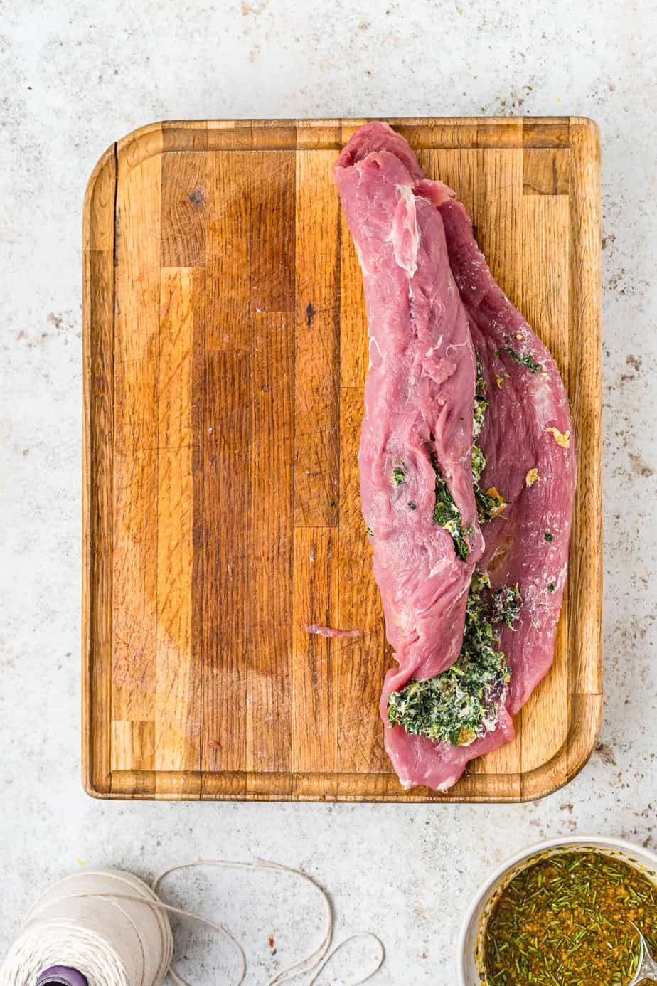 Pork tenderloin stuffed with spinach and cheese filling being rolled the longer direction on a cutting board on a kitchen counter from overhead with butchers twine nearby.