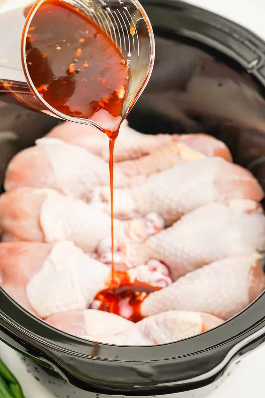 A slowcoooker full of raw chicken legs from the side with a hand pouring a sauce over the chicken from a bowl above.