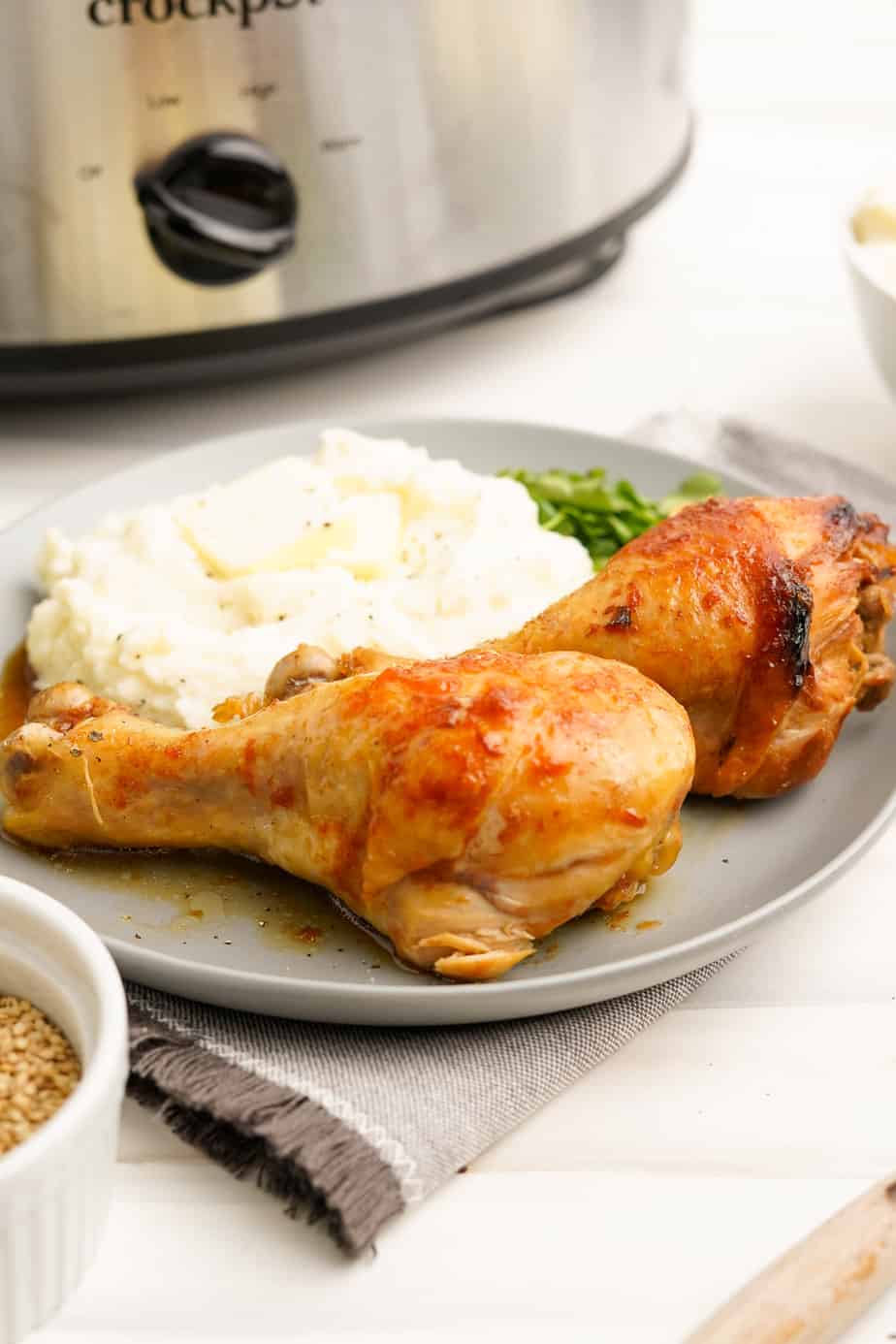 Two chicken drumsticks on a plate with mashed potatoes and a green vegetable from the side. A slowcooker sits behind the plate.