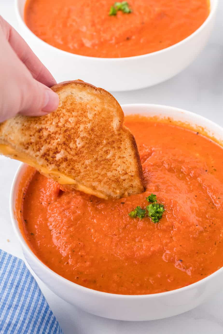 Dipping a grilled cheese sandwich into a bowl of tomato soup from the side with a second bowl in the background.