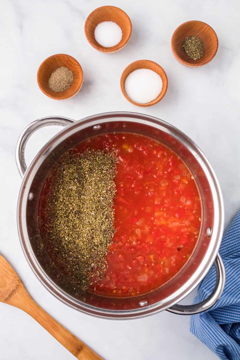 Italian herbs in a pot full of tomatoes from overhead with more herbs and spices in small bowls on the counter nearby.