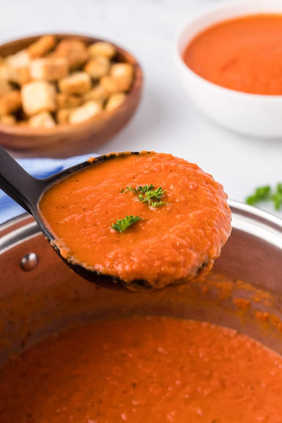 A ladle full of tomato soup from the side being lifted from the pot with a bowl of tomato soup and a bowl of croutons in the background.