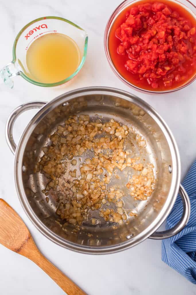 Cooking garlic and onion in a pan from overhead with a bowl of tomatoes and a measuring cup of chicken broth on the table nearby.