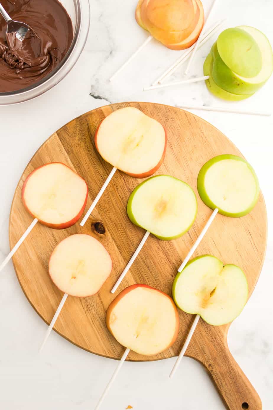 Fresh slices of apple cut into rounds with sticks added from the bottom on a cutting board from overhead on a counter with melted chocolate and more apples nearby.
