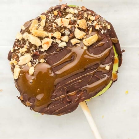 Close up of a slice of apple laying on parchment paper on a stick covered in caramel, chocolate and peanuts.
