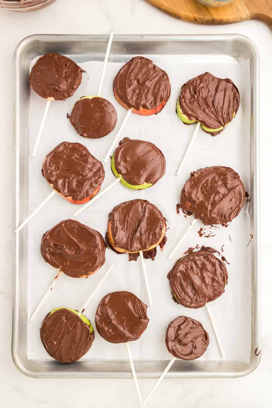 Apple slices with sticks coated in chocolate resting on parchment paper on a baking sheet from overhead.