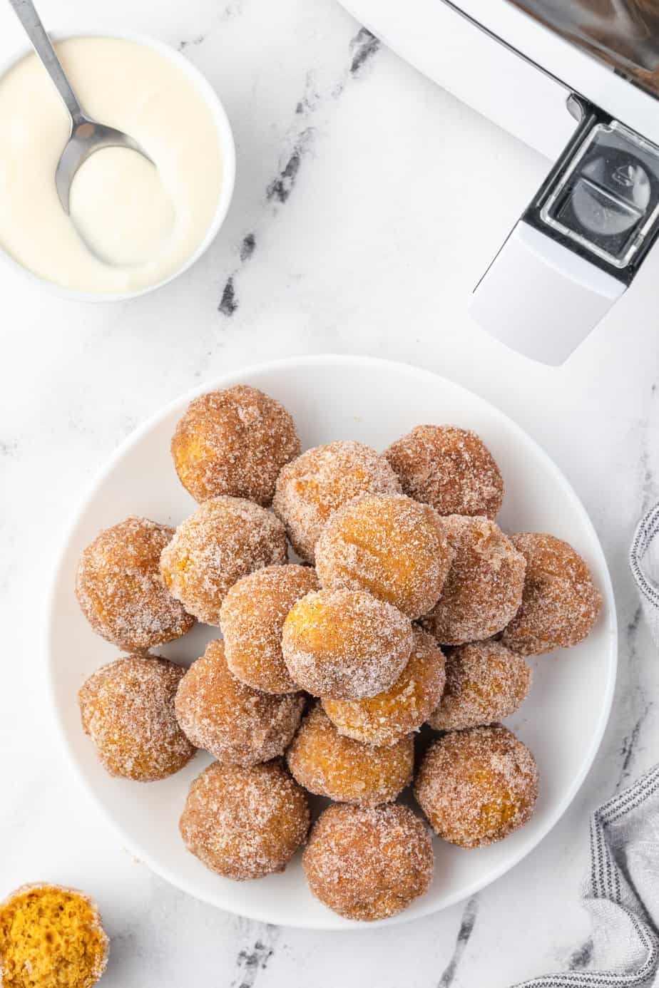 Pumpkin donuts on a large platter from overhead on a counter with cream cheese frosting with a spoon and an air fryer nearby.