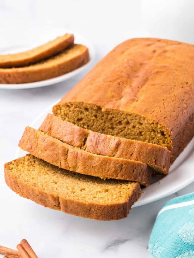 Pumpkin bread sliced at the end of a loaf resting on a plate with more slices of pumpkin bread in the background from the side on a counter.