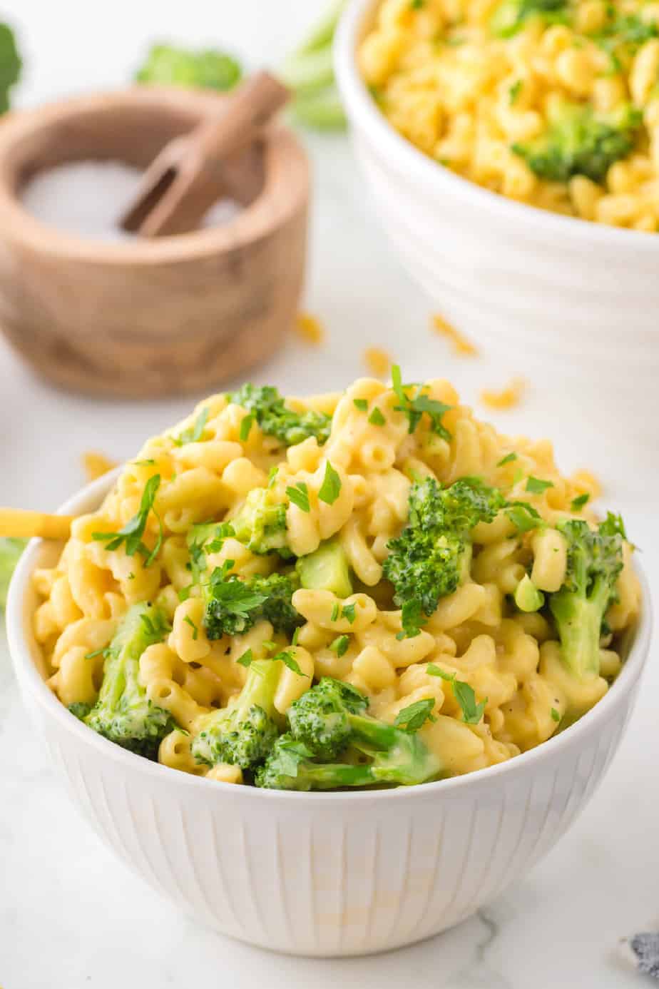 Macaroni and cheese with broccoli in a bowl on a counter with a serving bowl full of macaroni and cheese and a small bowl of salt in the background on the counter from behind.