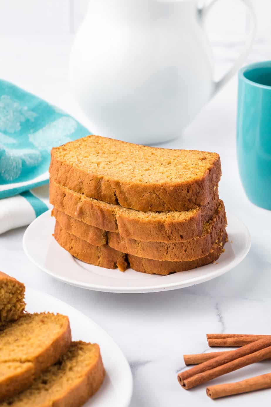 Pumpkin bread stacked on a plate on a counter from the side with a mug, pitcher, and more pumpkin bread on a platter along the edges.