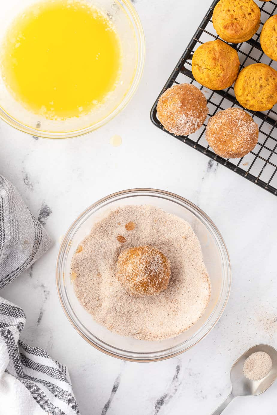 Rolling a pumpkin donut in a bowl of cinnamon sugar from overhead with a bowl of melted butter and more pumpkin donuts on a wire rack nearby on the counter from overhead.