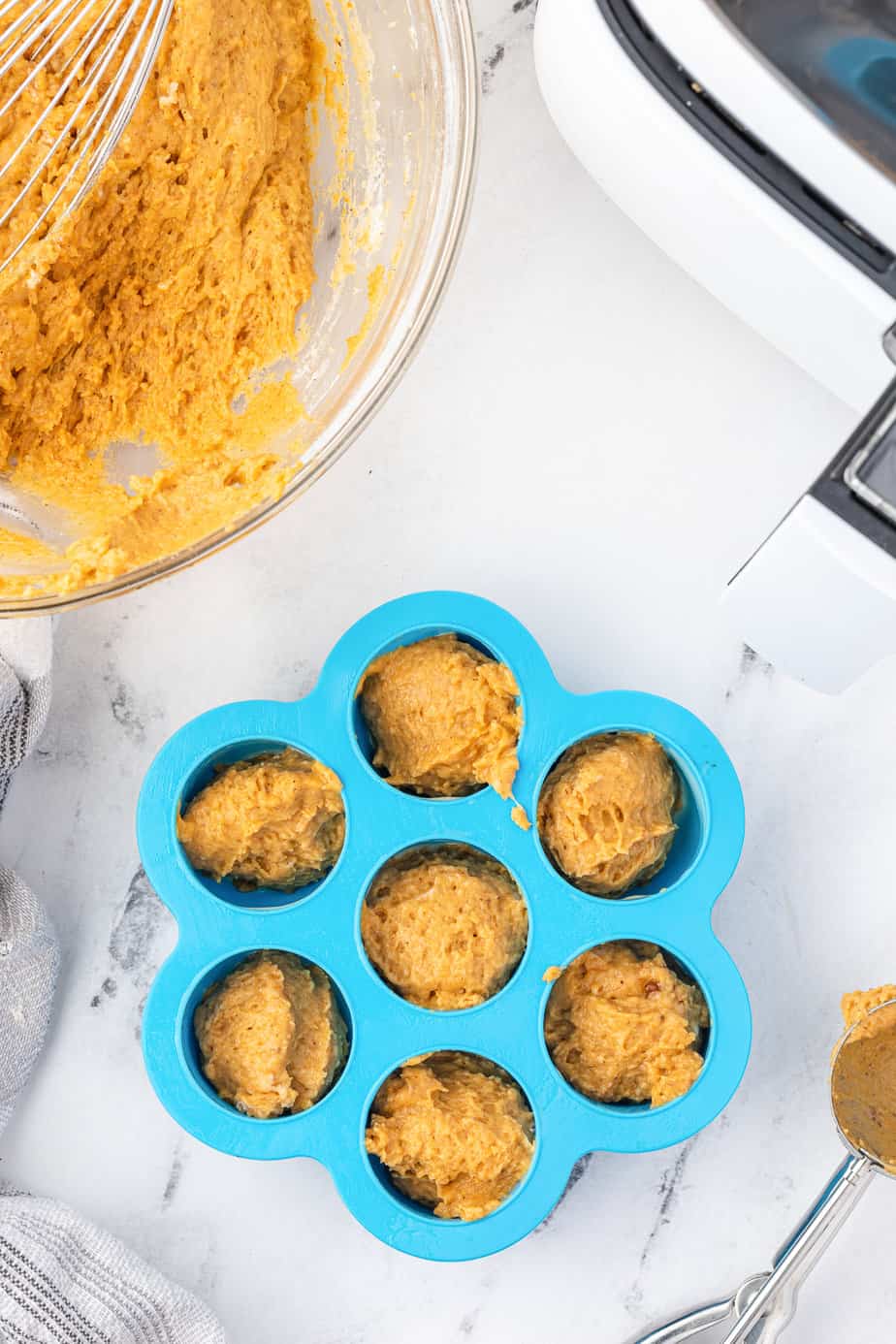 Pumpkin donut hole bites in a silicone mini muffin pan from overhead on a counter with a bowl full of more batter and an air fryer nearby.