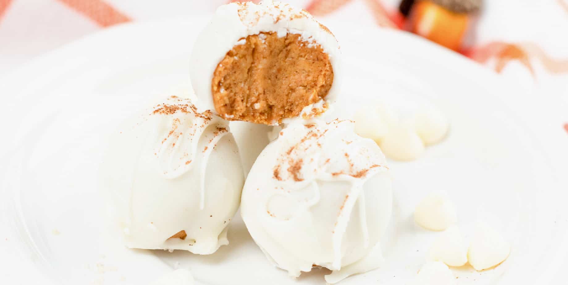 Wide view from the side of white chocolate coated pumpkin cheesecake bites on a white plate with the top piece missing a bite to show the pumpkin inside.