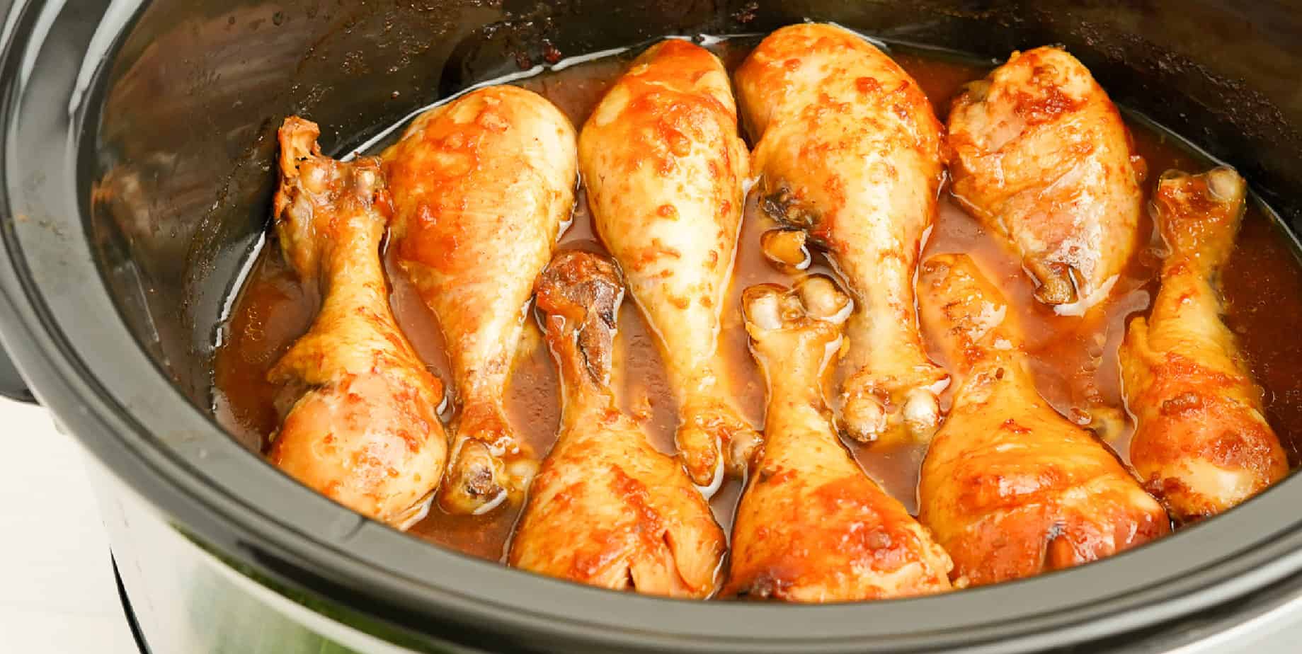 Chicken drumsticks lined up in the base of a slow cooker covered in sauce.