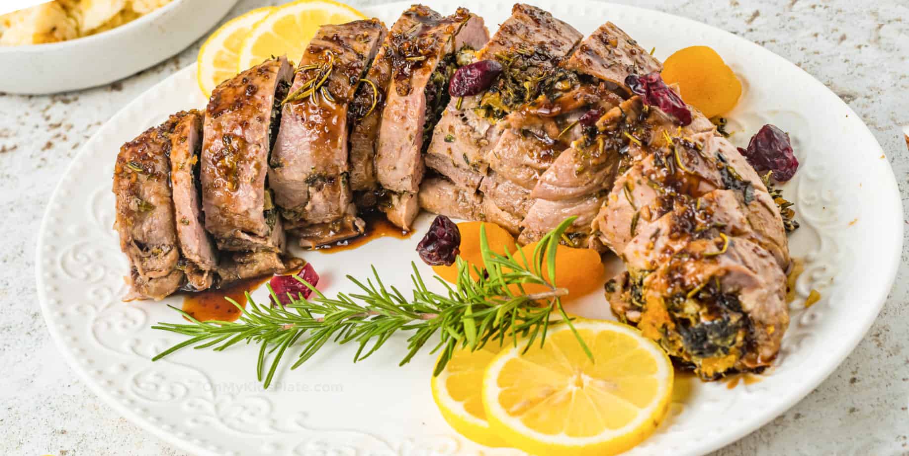 Stuffed sliced pork tenderloin on a oval platter garnished with fresh rosemary, dried fruit and lemon on a counter from the side.