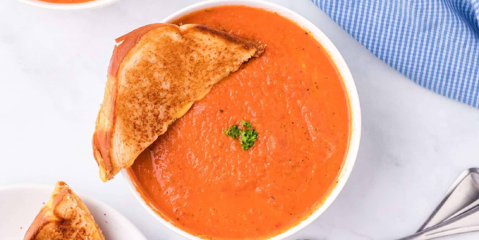 Wide view of a bowl of tomato soup with half a grilled cheese sandwich dipping in from overhead.