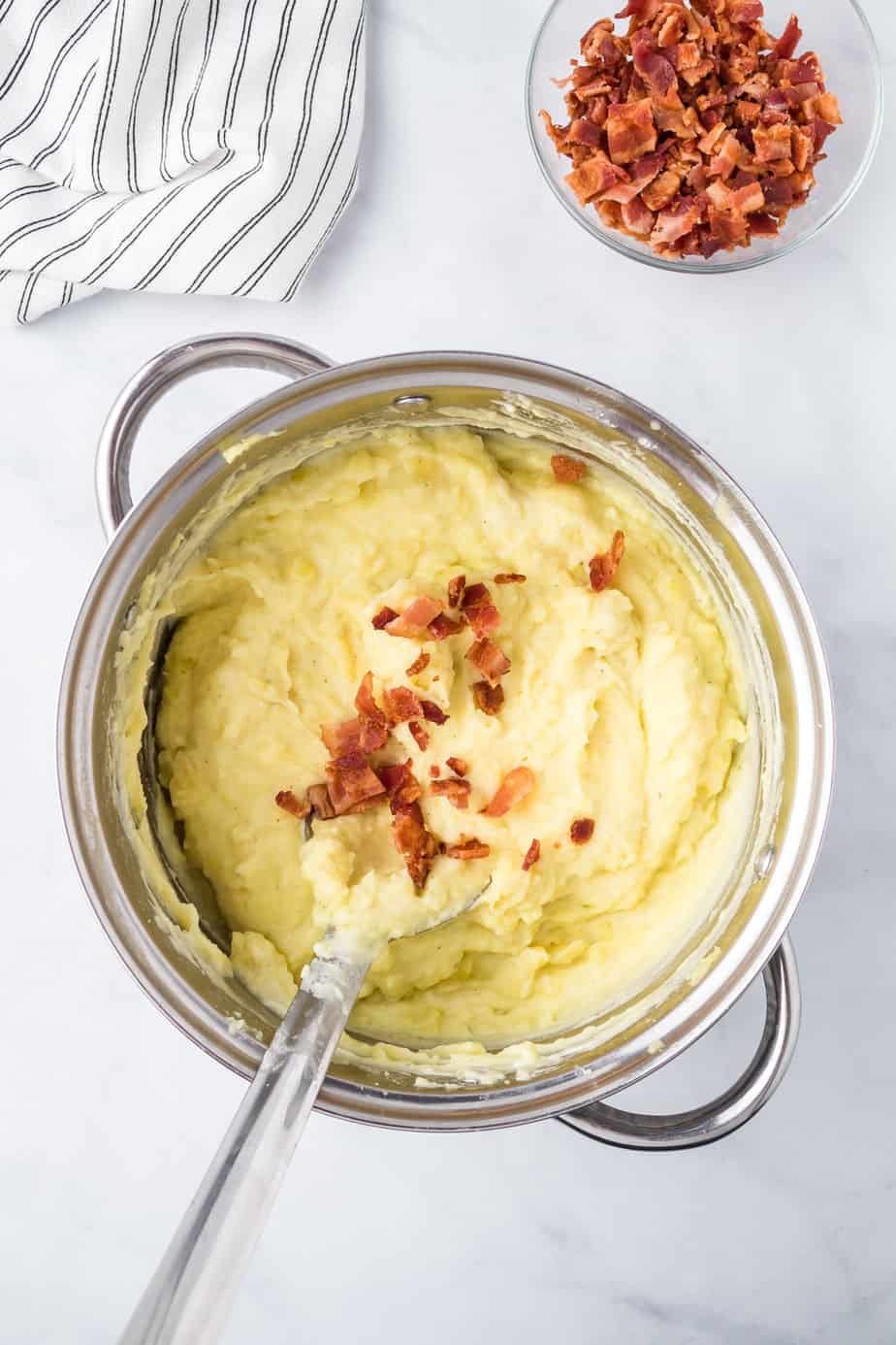 Adding chopped bacon to smooth mashed potatoes in a large pot with a spoon from above on a counter.